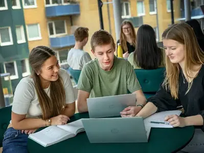 Tre elever samarbetar framför sina bärbara datorer, med böcker och anteckningar framför sig.