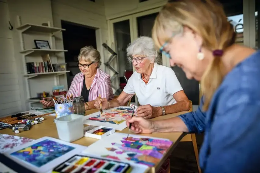 Tre kvinnor sitter och målar med akvarellfärger runt ett bord